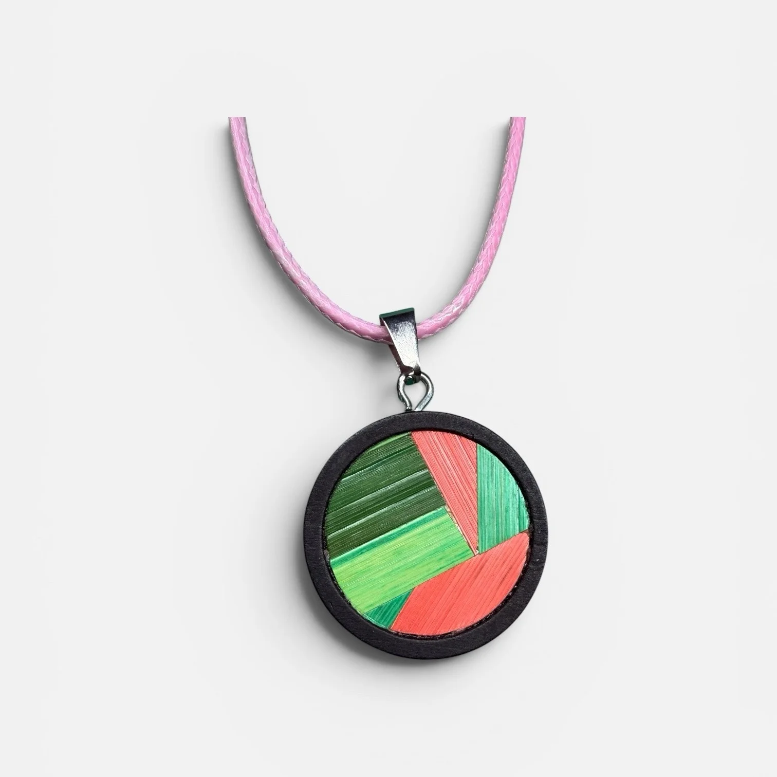 Collier médaillon vert — médaillon rond cerclé de bois noir, marqueterie de paille de seigle en camaïeu de verts et corail, cordon rose, fond blanc