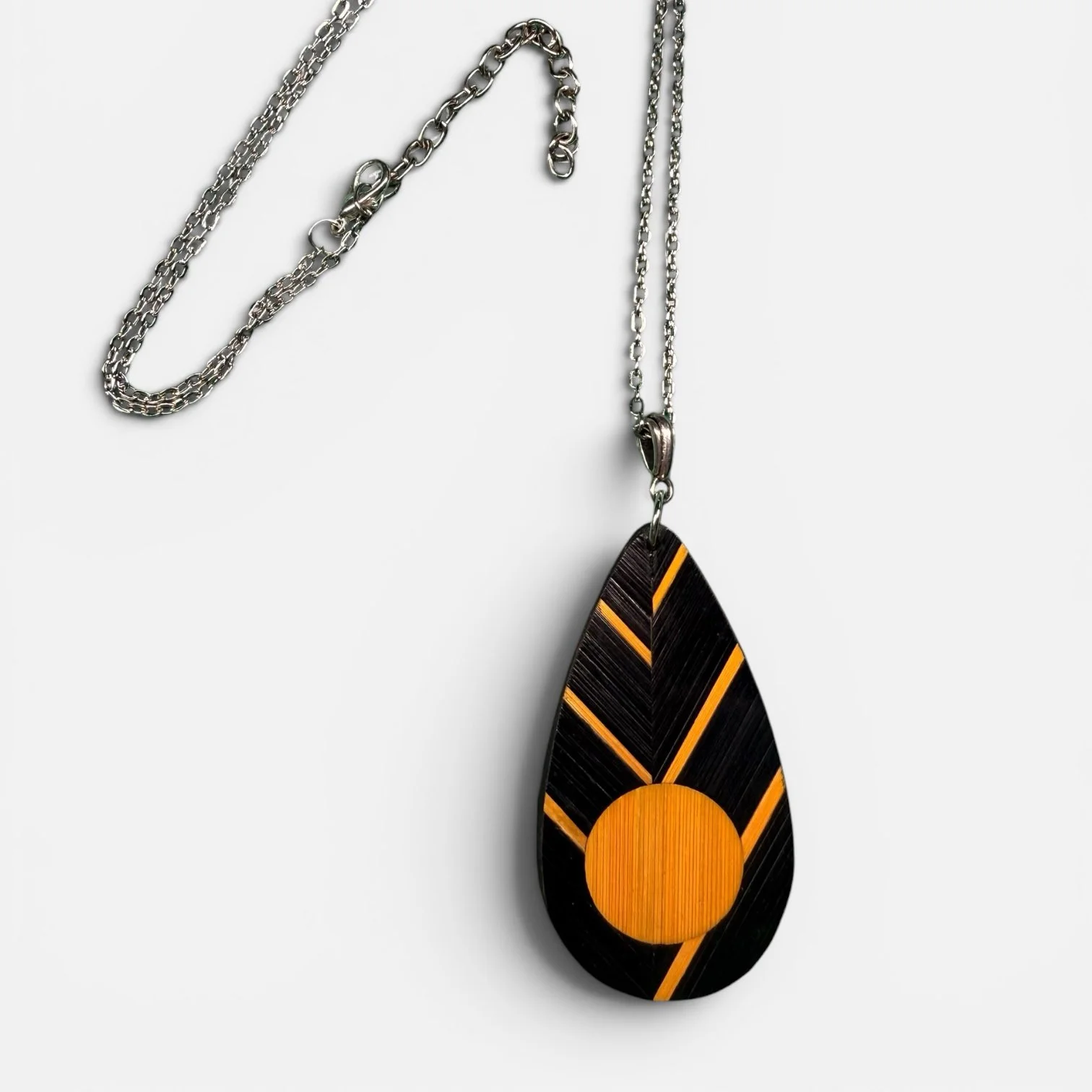 Collier sautoir Abeille — pendentif goutte en marqueterie de paille de seigle teintée noir et orange, disque et lignes rayonnantes, chaîne acier inoxydable argenté, fond blanc